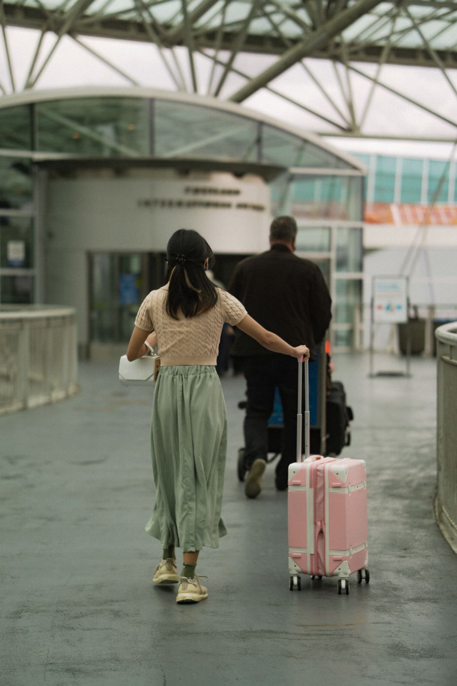 woman in brown t shirt pushing her pink luggage and man in black long sleeves pushing trolley — Docs: ホームからホームへ：チケット、時刻表、タクシーが片手に。