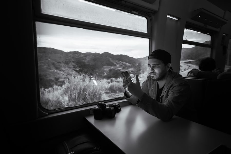 Man using a smartphone on a train journey through scenic landscape. — Developers: カフェオフィス:テザリング稼働、ビデオ通話は安定、ホテルWi-Fiのロシアンルーレットなし。