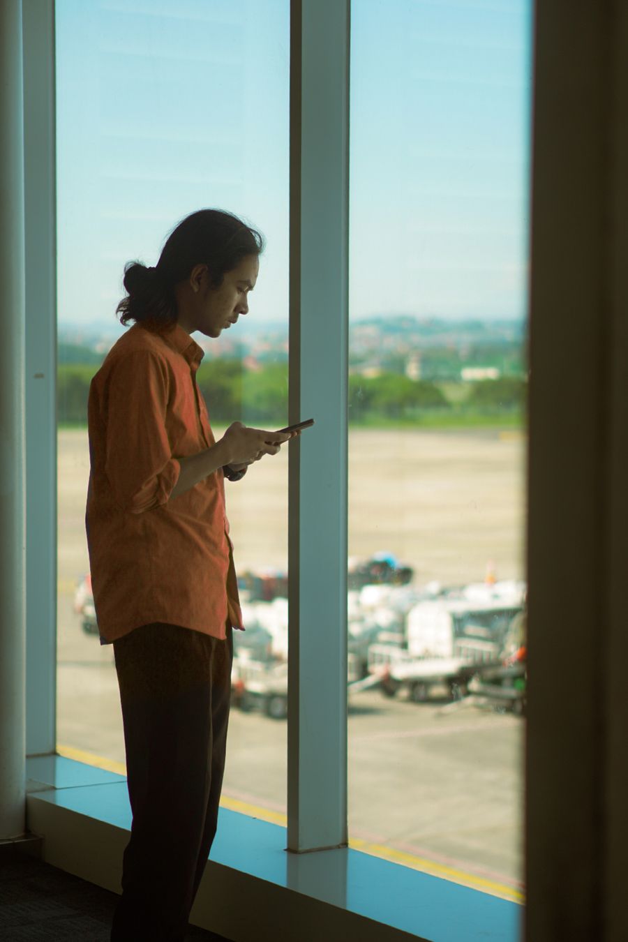 Man in orange shirt using a smartphone while standing at an airport window, overlooking the runway. — Old town, new tricks: translation apps working in real time.