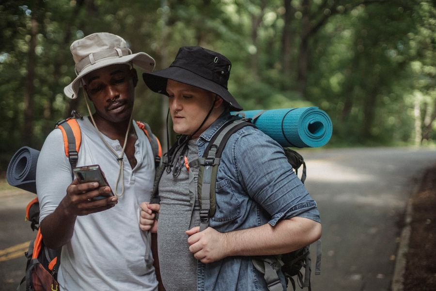 Young multiethnic tourists with hiking equipment surfing internet on cellphone on roadway against trees. — Market mornings: look up the dish, translate the menu, pay by phone.